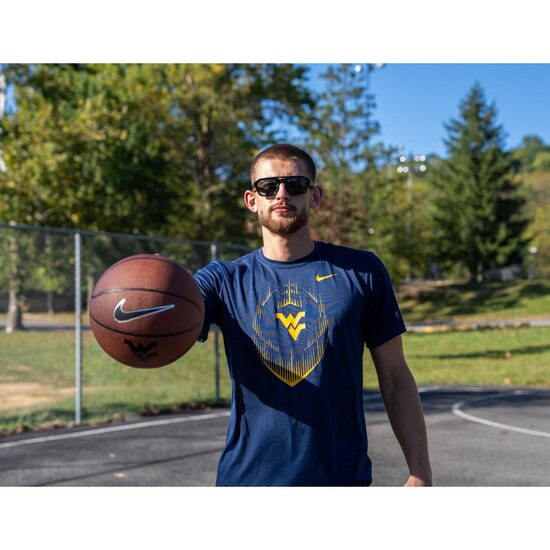 Erik Stevenson holding out a WVU basketball while sporting our navy Nike WVU short sleeve shirt with football icon and Flying WV as chest graphic